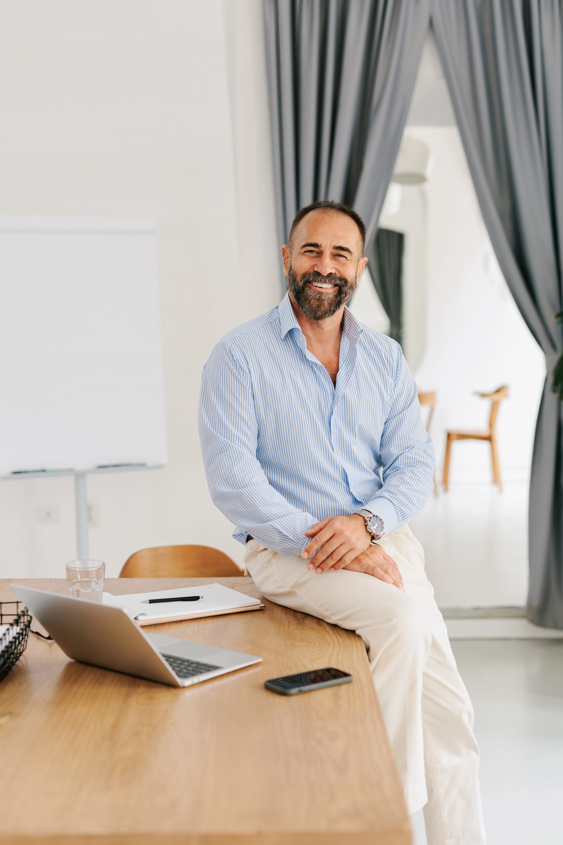 A businessman leaning against his desk – on which sit a laptop and notebook – smiling and looking at the camera.