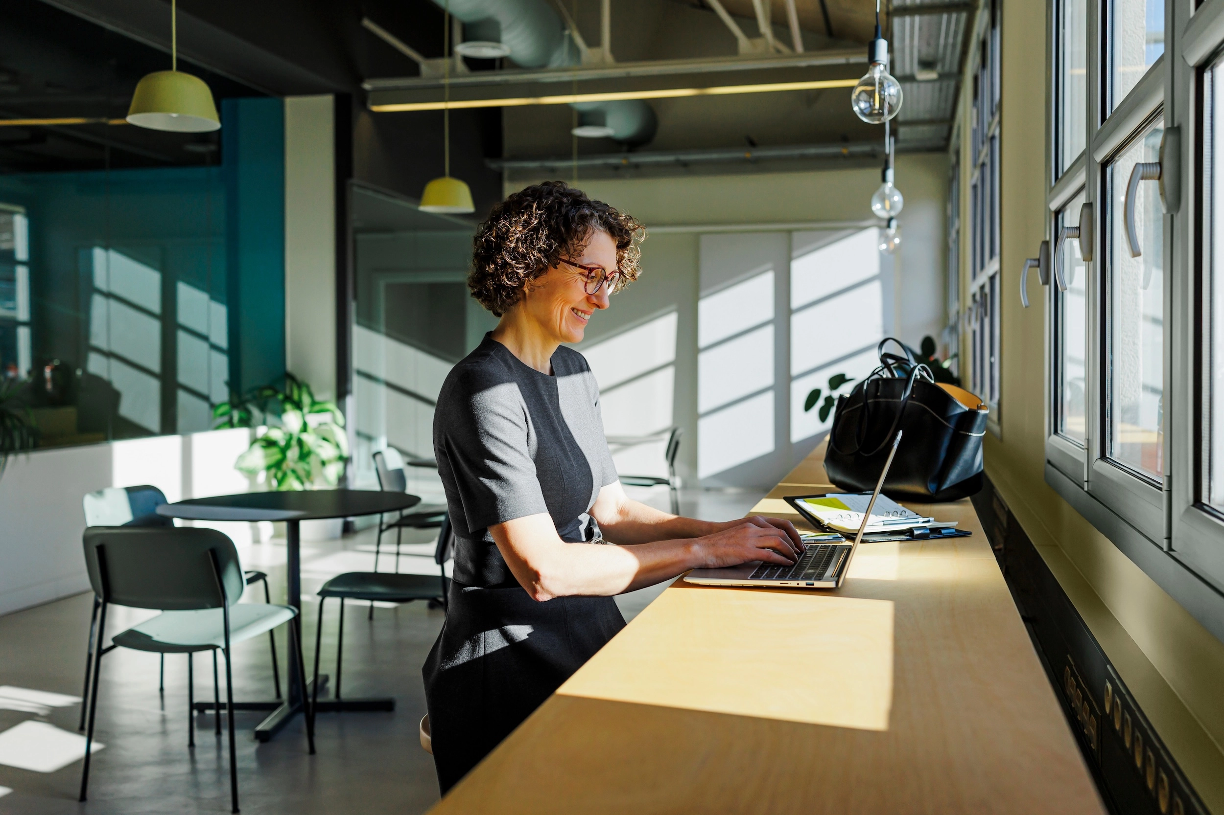 A woman sitting at a counter in a co-working space, smiling, typing on her laptop.
