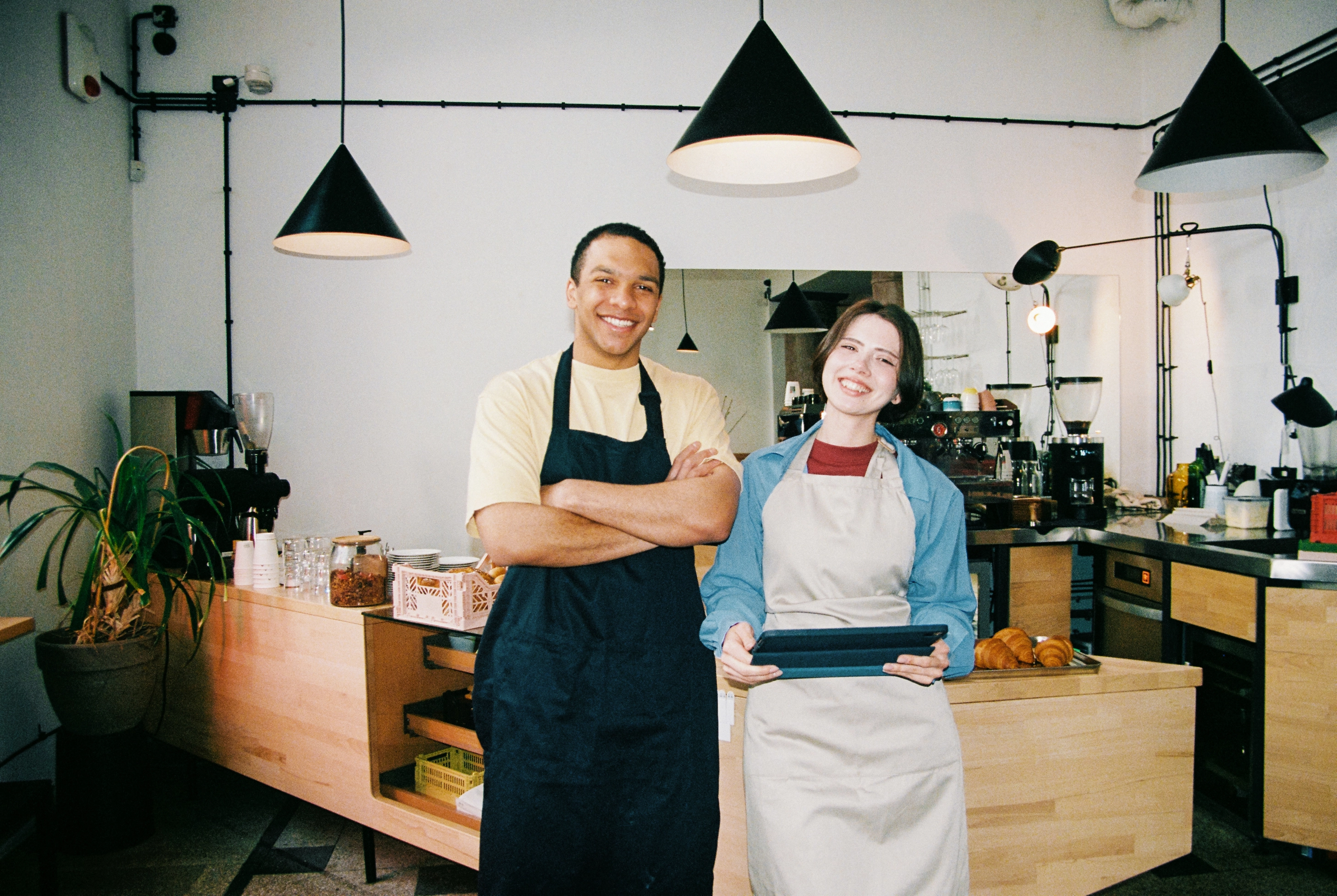 Two coffee shop workers posing in front of the counter. One is holding a tablet.
