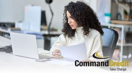 A woman sitting at an office desk, holding a pen and paper, looking at a computer screen.